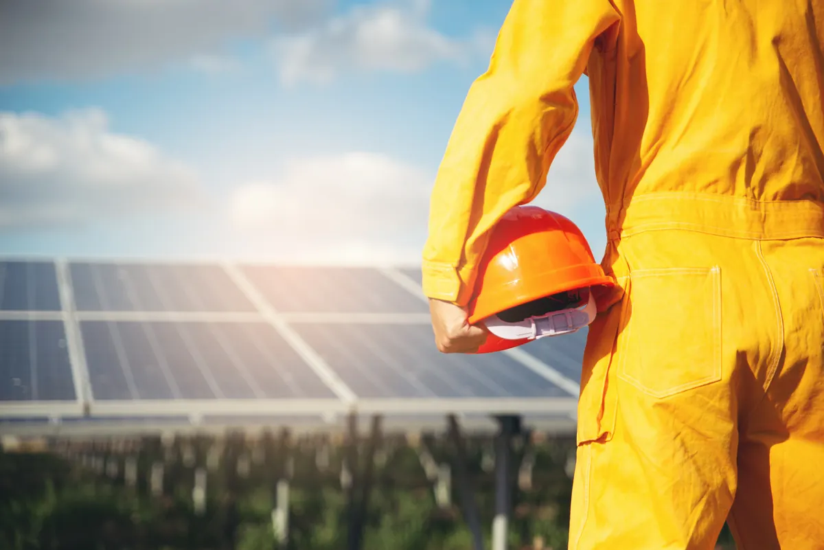 Worker in yellow coveralls holding a hard hat beside a row of solar panels