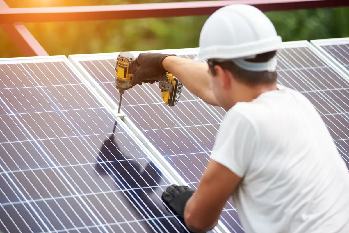 Worker in a hard hat fastening a solar panel with a drill